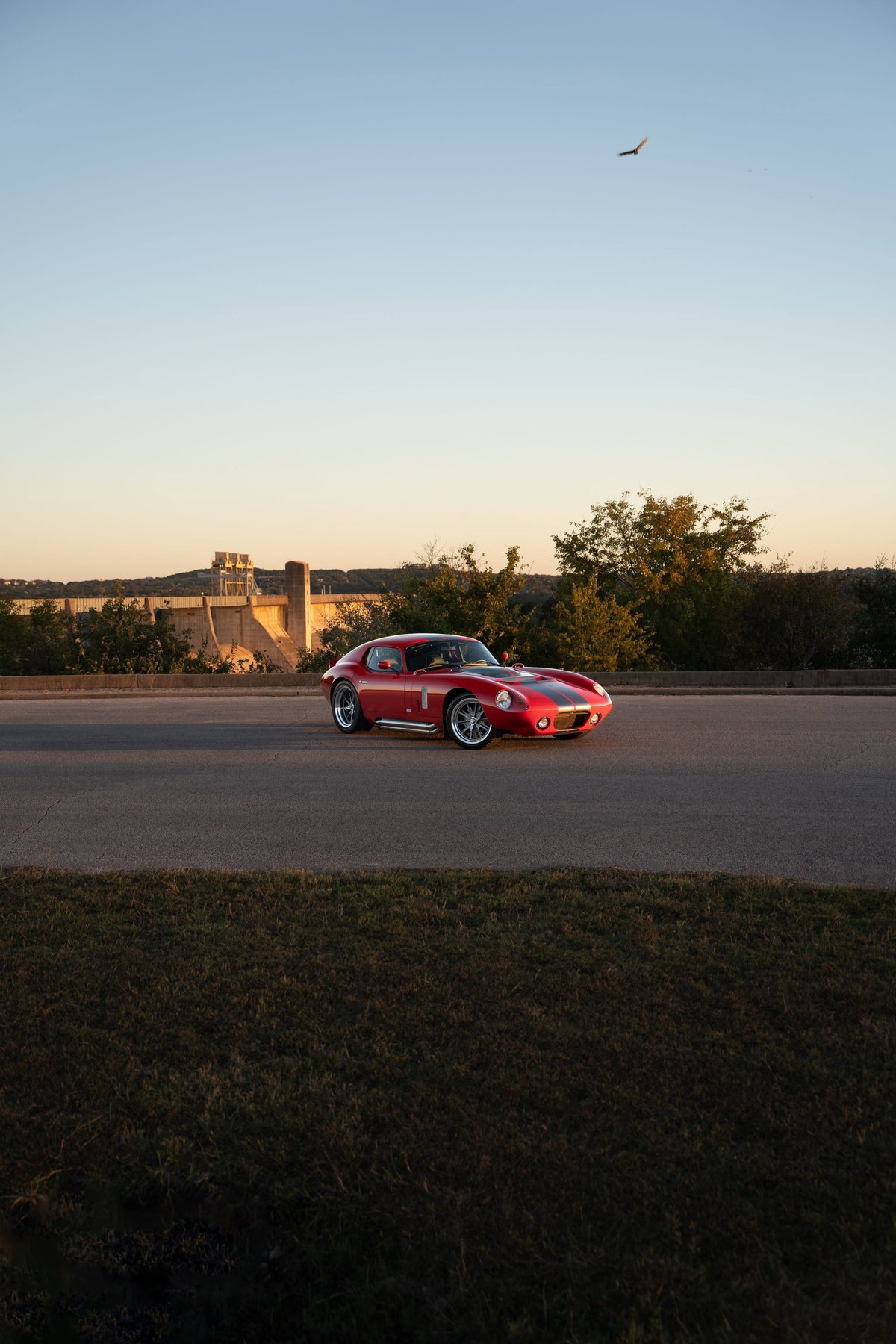 Red 1965 Superformance Shelby Daytona CSX with black racing stripes and chrome rims drives past a dam.