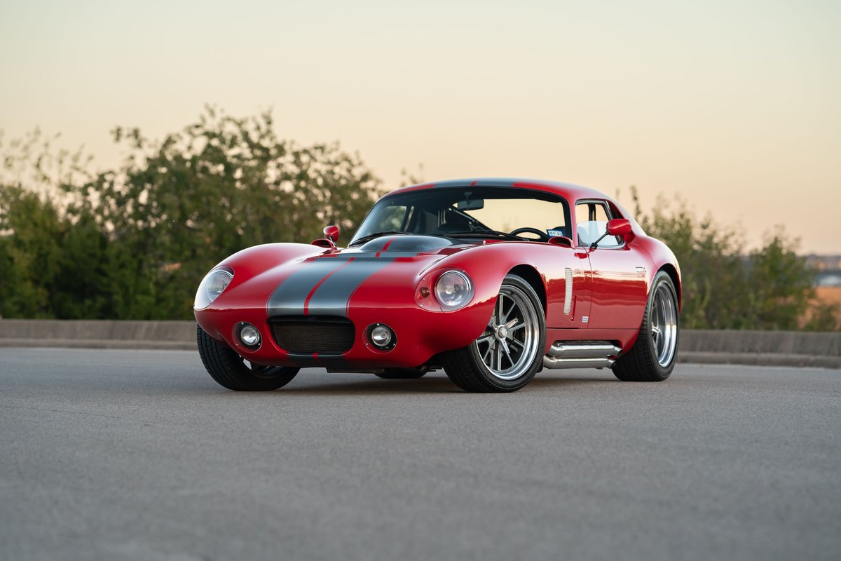 Red 1965 Superformance Shelby Daytona CSX coupe with dual gray racing stripes, chrome side pipes, and alloy rims.