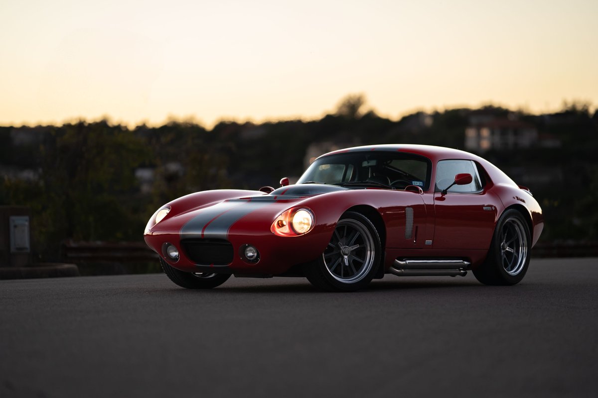 Red 1965 Superformance Shelby Daytona CSX with dual gray racing stripes and chrome side pipes.