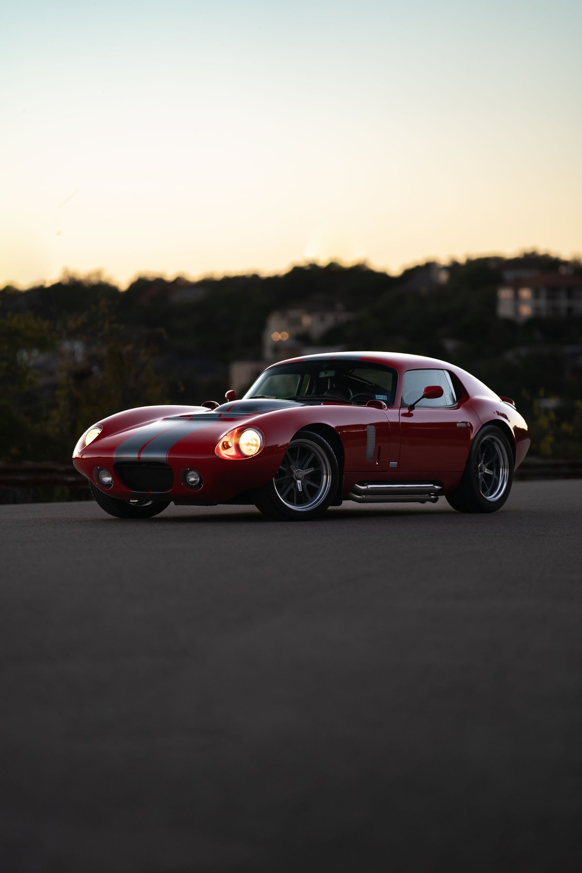 A red 1965 Superformance Shelby Daytona Coupe with gray racing stripes, custom rims, and side exhaust.