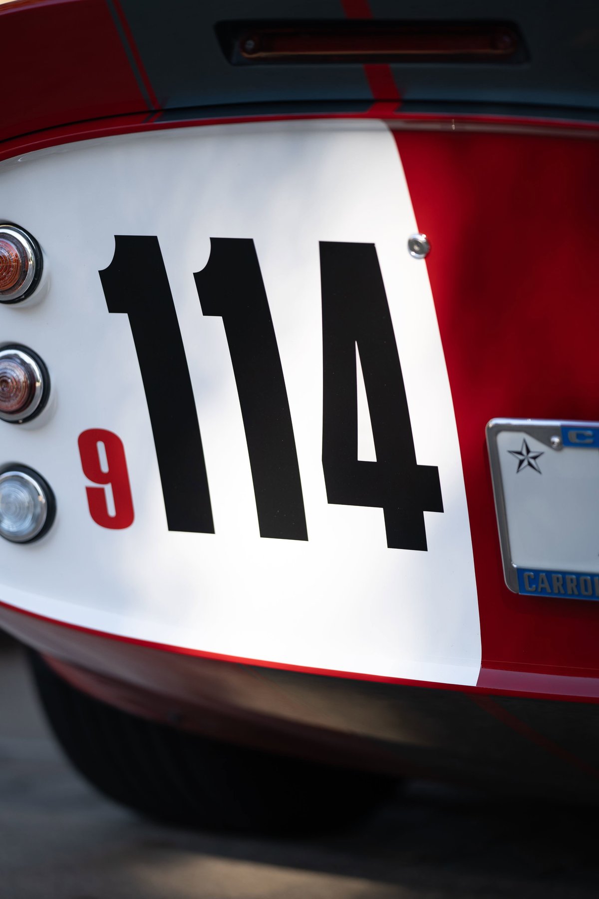 Rear close-up of a red 1965 Shelby Daytona CSX with a white stripe and race numbers 114 & 9.