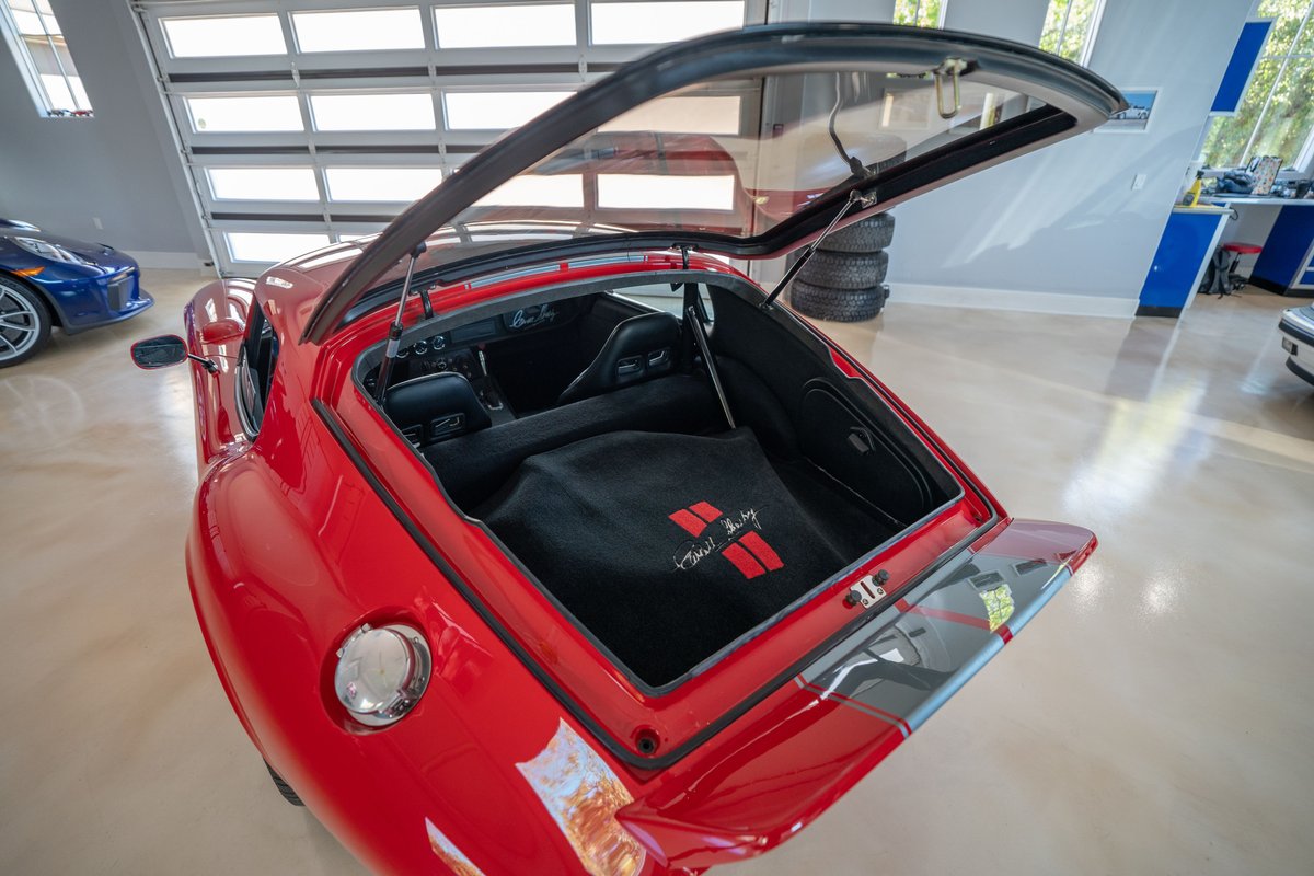 Red 1965 Shelby Daytona Coupe with trunk open, showing black interior carpet with red racing stripes.