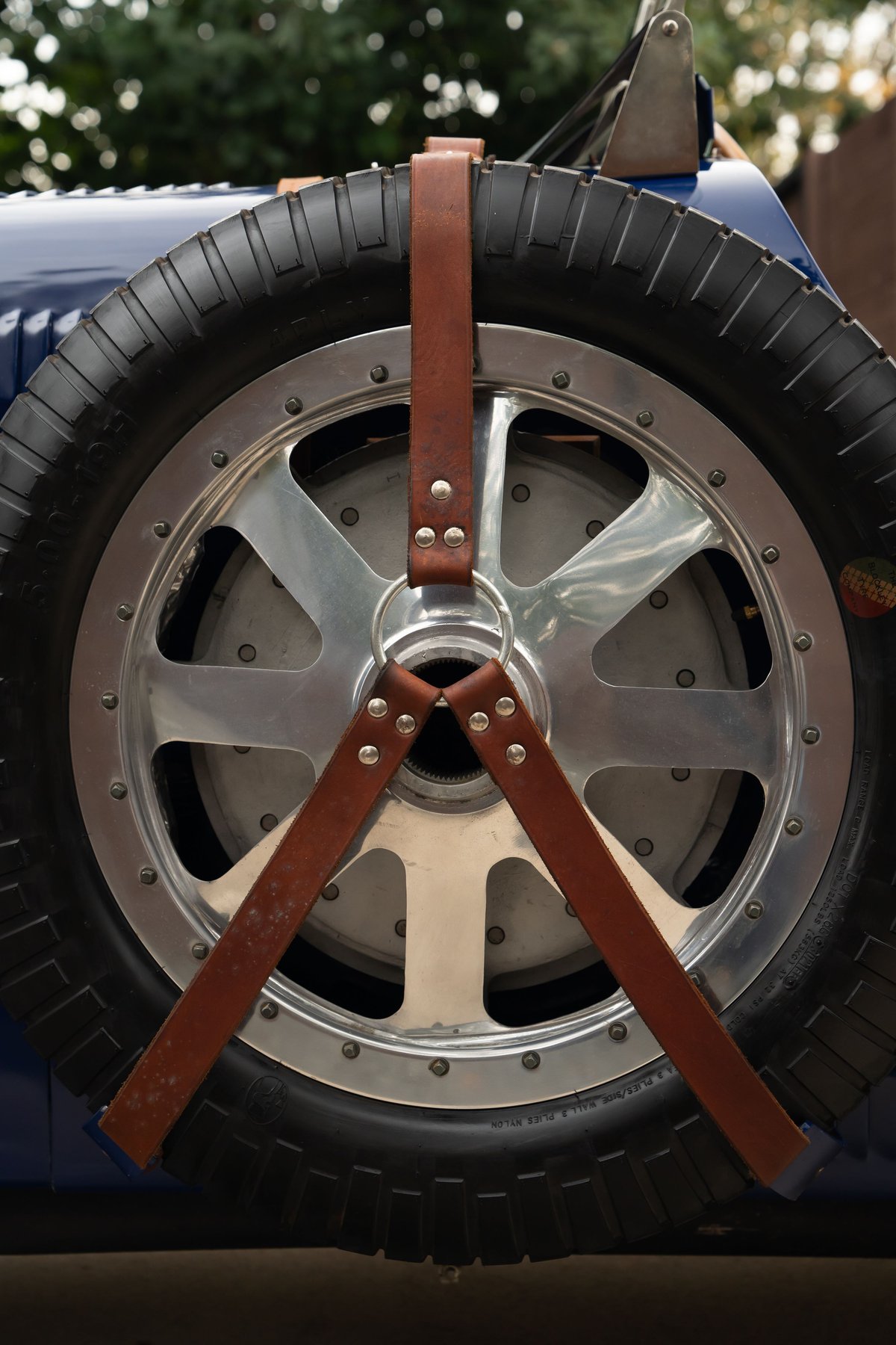 Close-up of a spare tire on a blue 1927 Bugatti Type 35. The polished metal wheel features a three-strap leather tie-down.