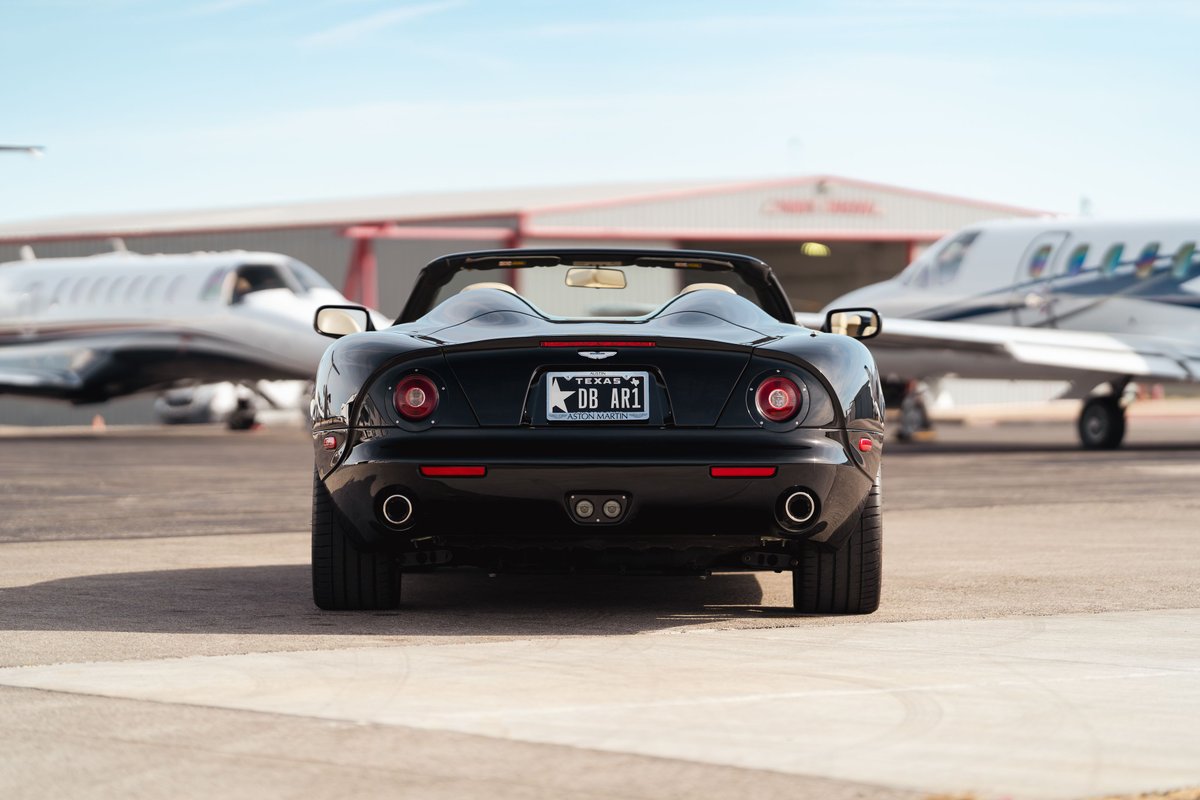 Rear view of a black 2003 Aston Martin DB AR1 Zagato convertible, parked on tarmac by private jets.