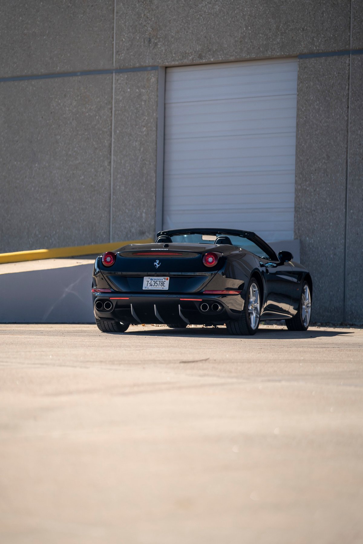 Black 2016 Ferrari California T convertible with red trim and silver rims, parked outside a building.
