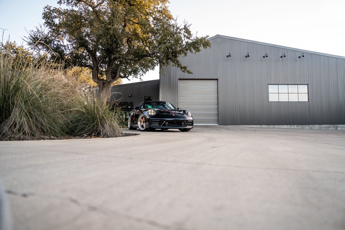 Black 2023 Porsche 911 Carrera 4 GTS with bronze rims parked outside a metal building.
