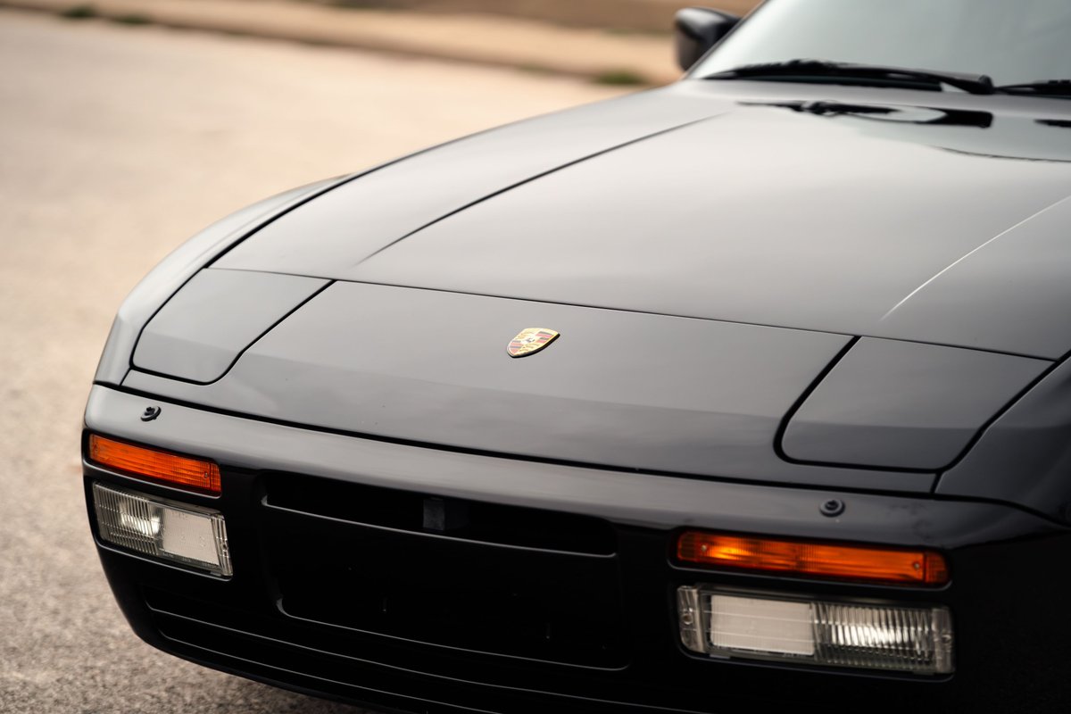 Close-up of the front end of a black 1986 Porsche 944 Turbo, showing pop-up headlights and a Porsche emblem.