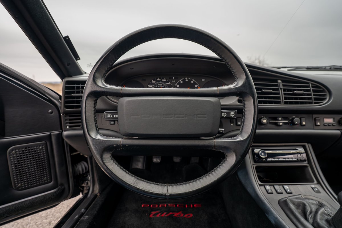 Dashboard of a black 1986 Porsche 944 Turbo, featuring a leather-wrapped steering wheel and "Porsche Turbo" floor mats.