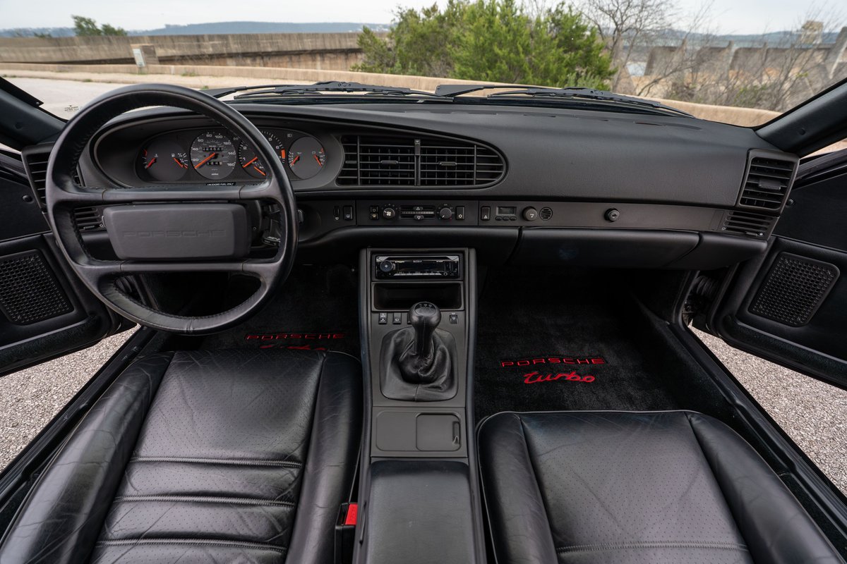 Interior view of a black 1986 Porsche 944 Turbo with leather seats and red "Porsche Turbo" floor mats.
