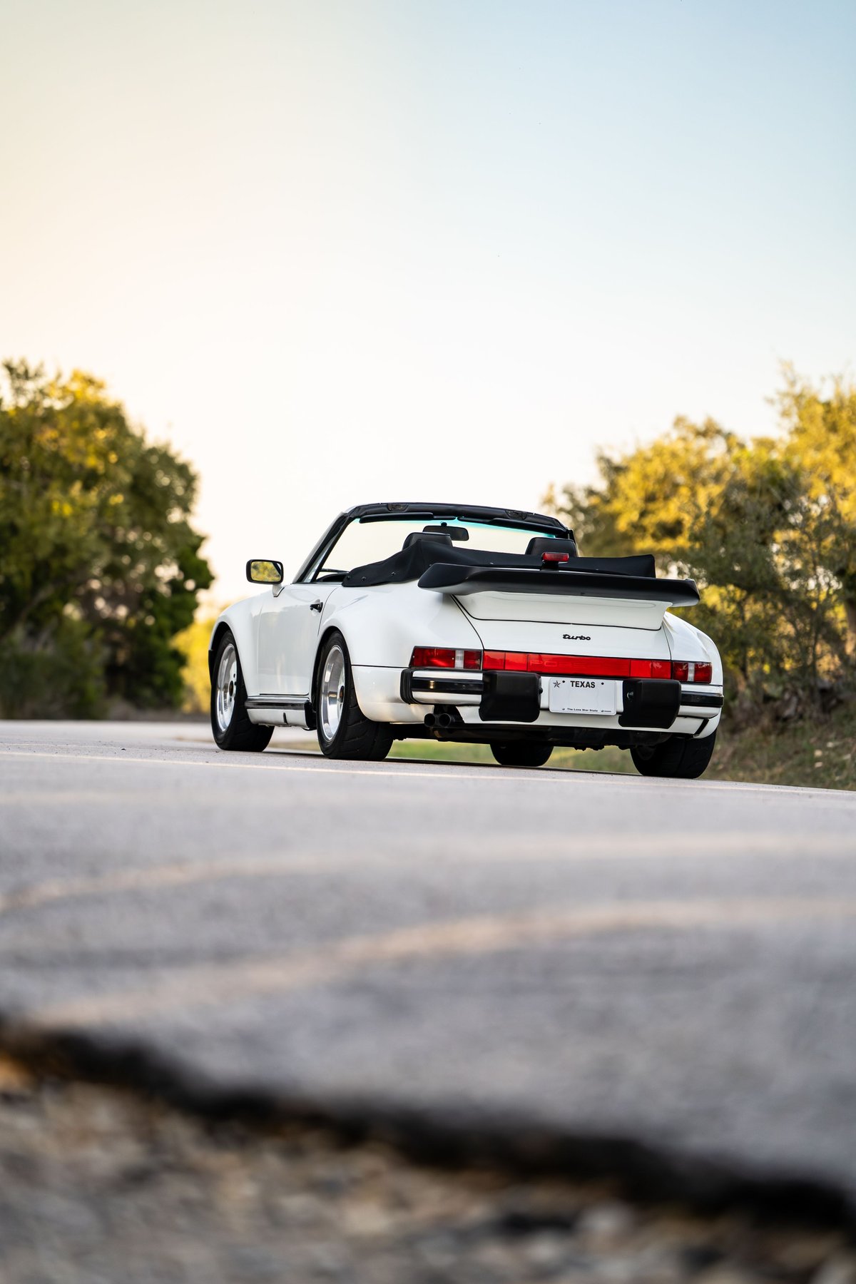 White 1988 Porsche 930 Turbo slantnose convertible with black soft top and whale tail spoiler.