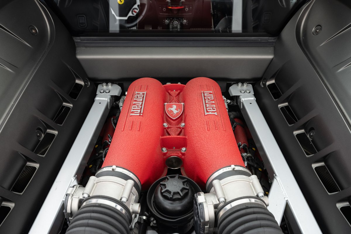 Close-up of a Ferrari F430 engine bay. The V8 engine features red intake runners with Ferrari badging.
