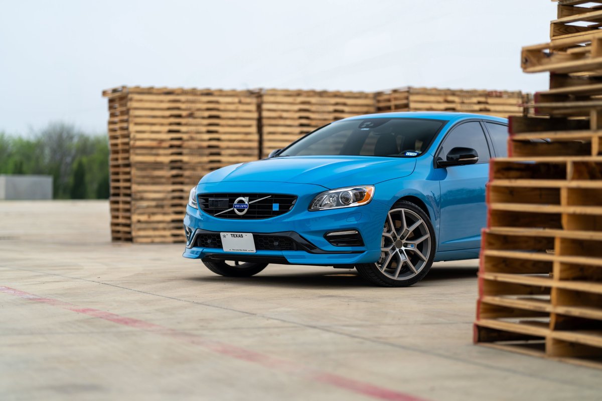 A bright blue 2015 Volvo V60 Polestar with unique silver rims sits in front of stacked wooden pallets.
