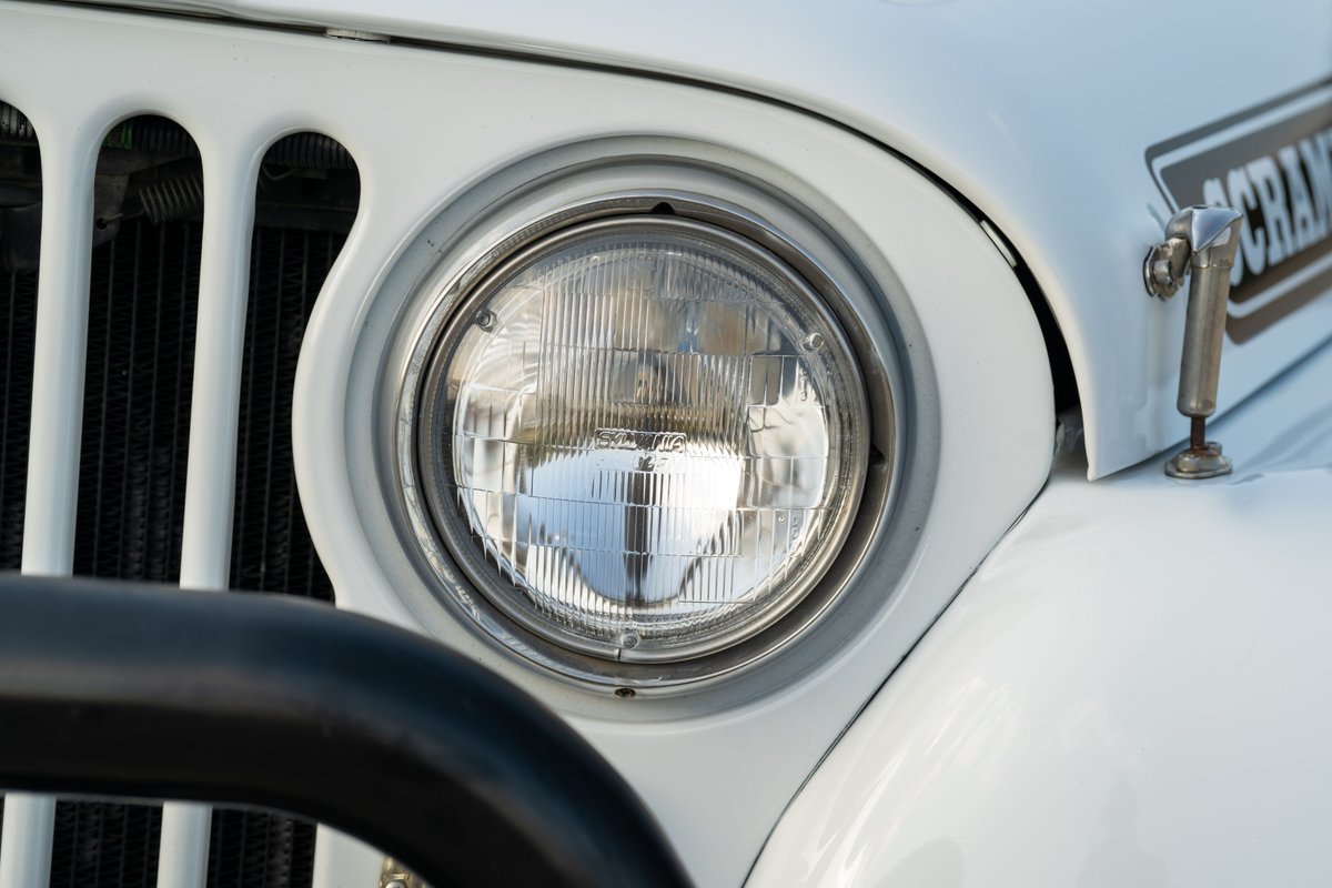 Close-up of a white 1983 Jeep Scrambler's round headlight, chrome trim, and grille.