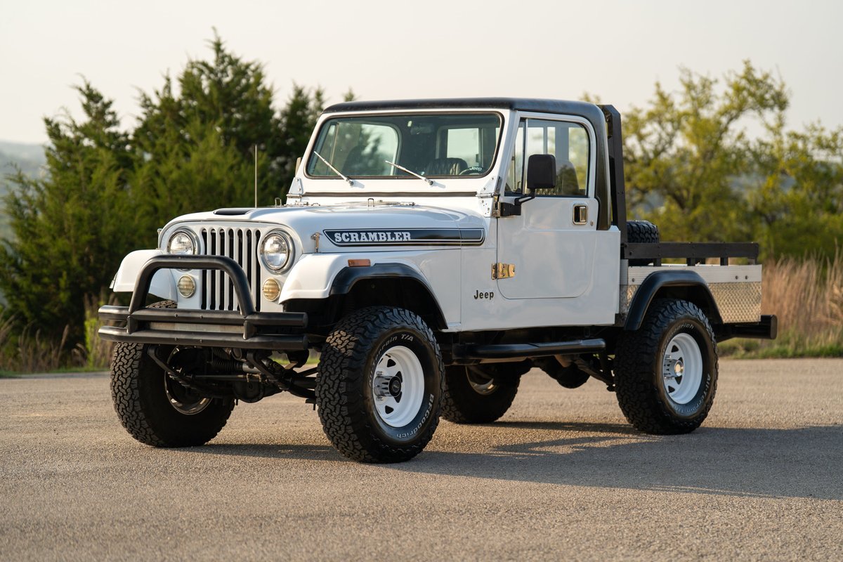 1983 white Jeep Scrambler CJ-8 flatbed pickup with white rims and aggressive tires.