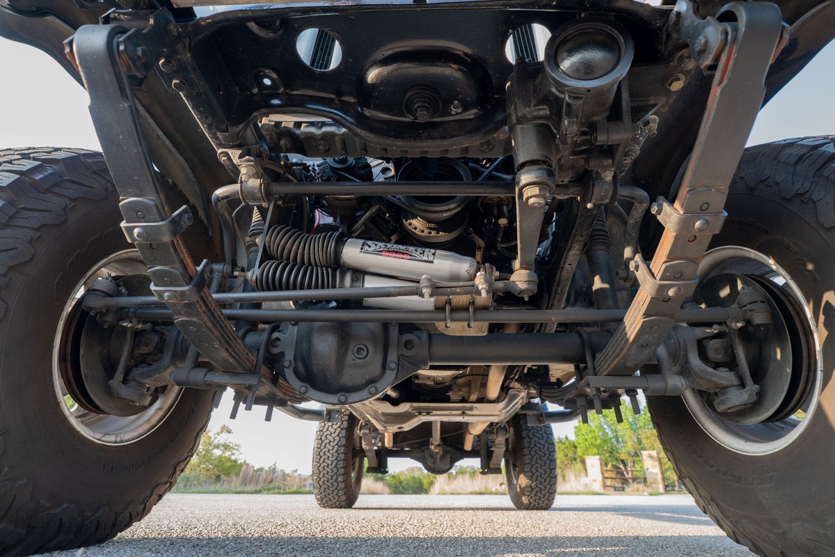 Underside view of a lifted white 1983 Jeep Scrambler showing leaf spring suspension & heavy-duty steering components.
