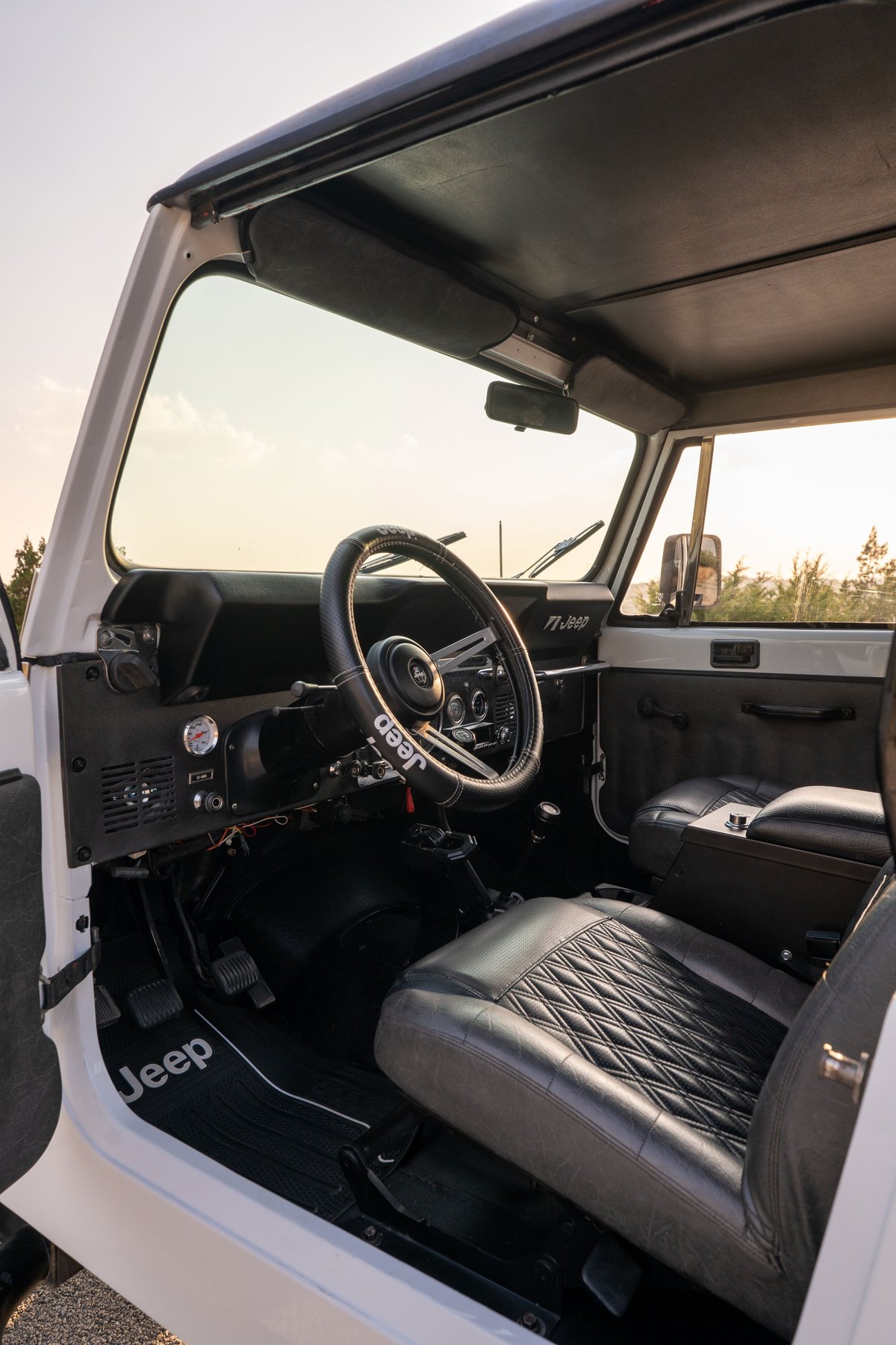 Interior of a white 1983 Jeep CJ8 Scrambler, showing diamond-stitched seats, Jeep floor mats, and a custom steering wheel.