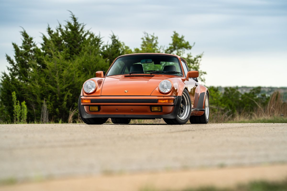 Low-angle view of an orange metallic 1986 Porsche 930 Turbo coupe with black accents and period-correct rims.