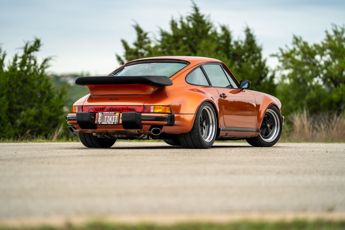 1986 orange metallic Porsche 930 Turbo with a whale tail spoiler and black trim.