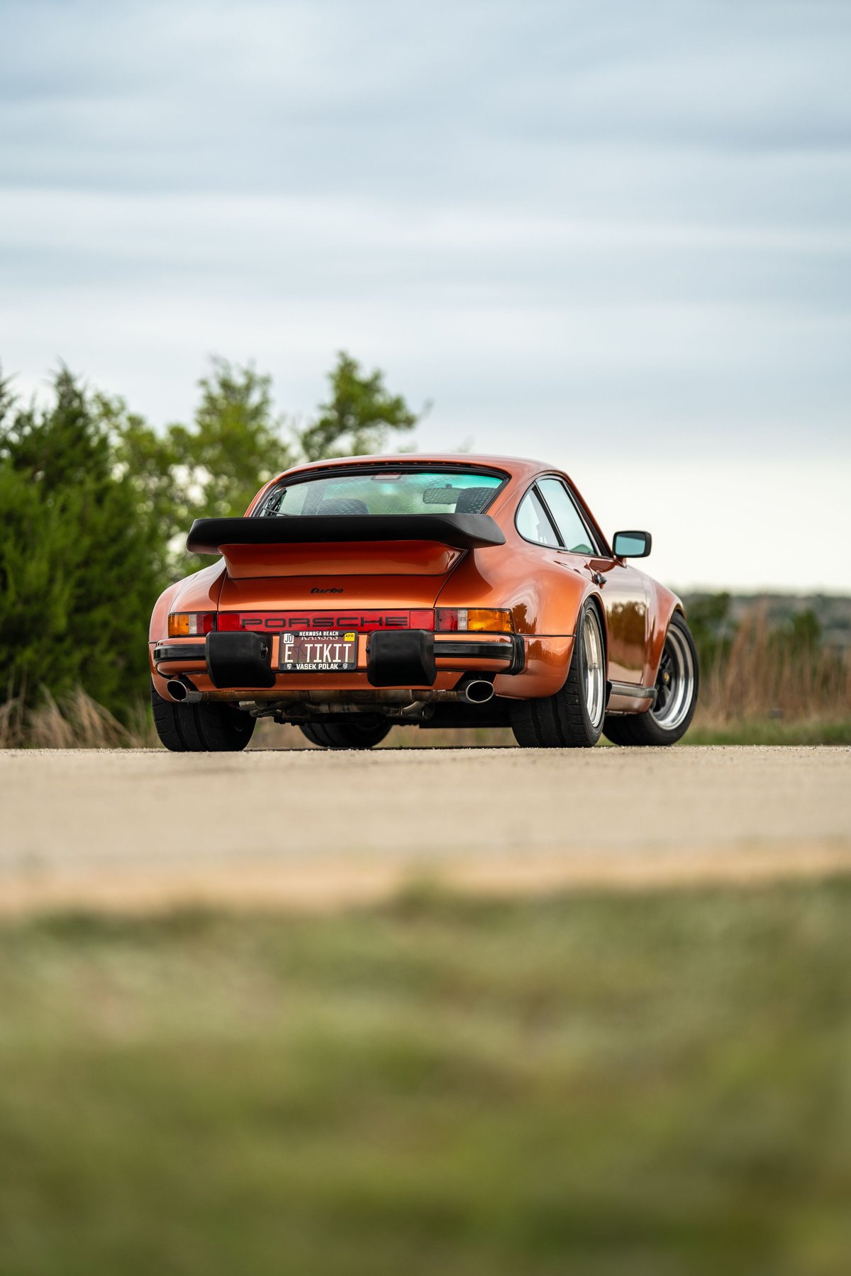 Rear view of an orange metallic 1986 Porsche 930 Turbo with a black whale tail spoiler and chrome rims.