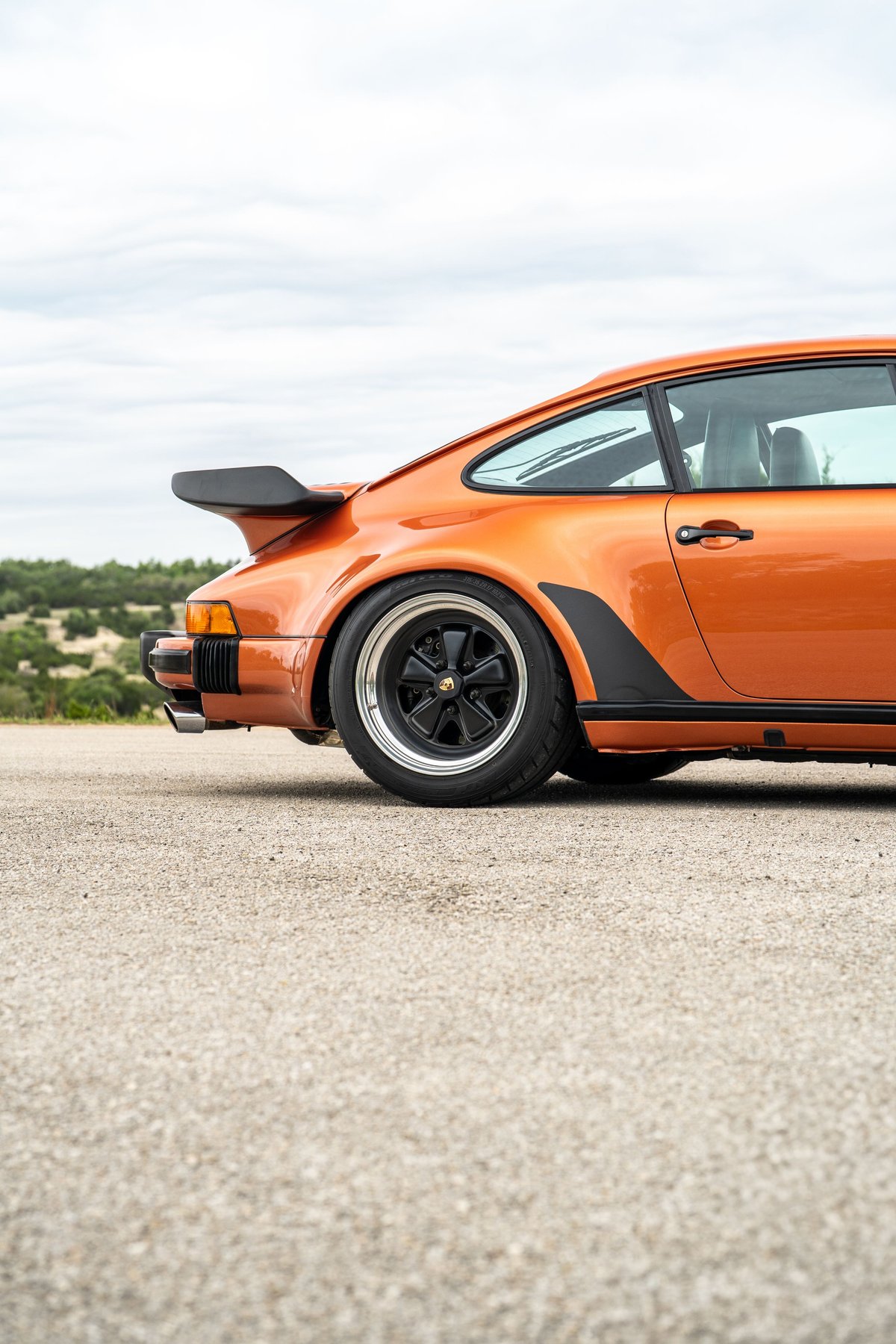1986 Porsche 930 Turbo in orange metallic, featuring a whale tail spoiler, black trim, and Fuchs rims.
