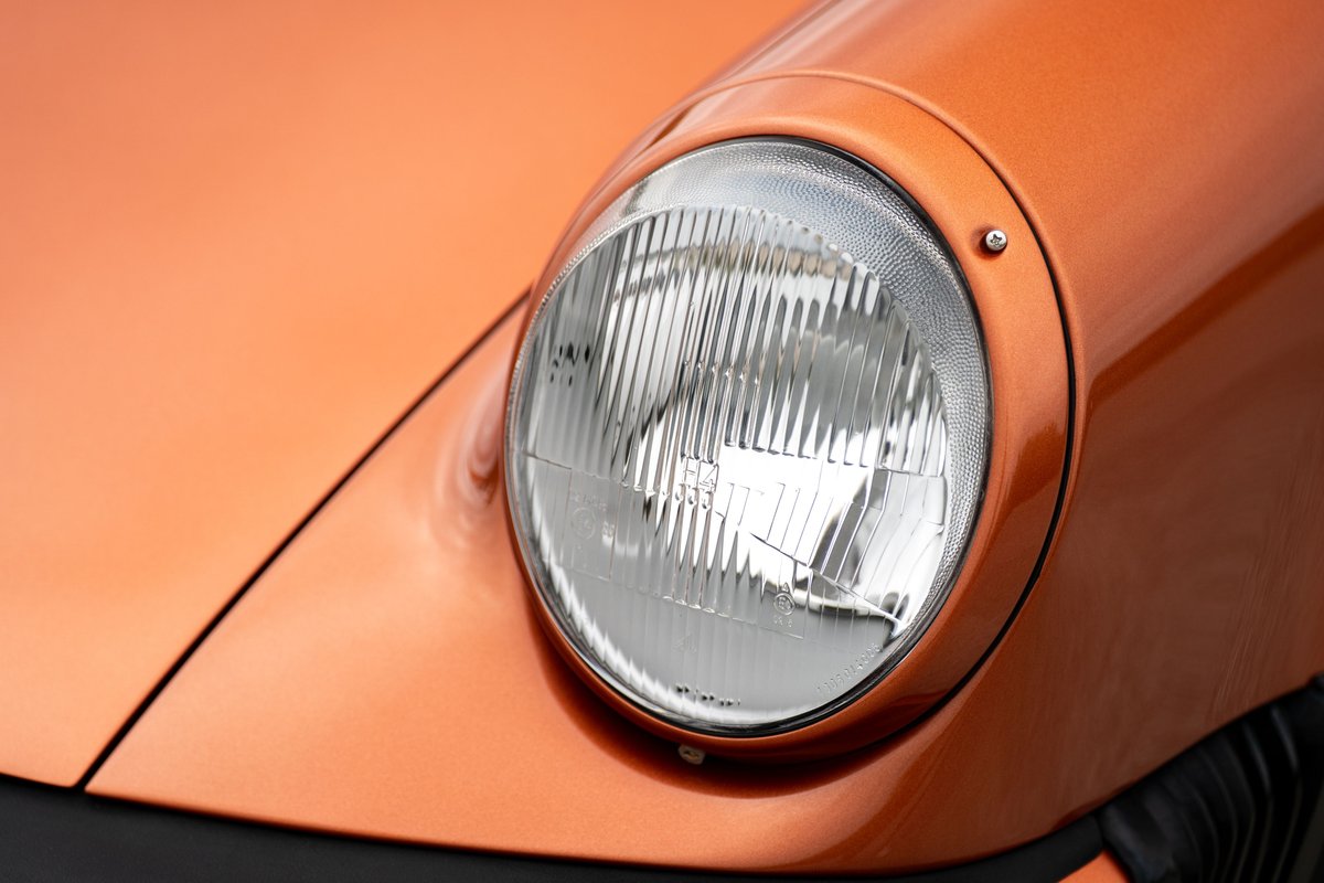 Close-up of a 1986 Porsche 930 Turbo's round headlight in orange metallic paint.