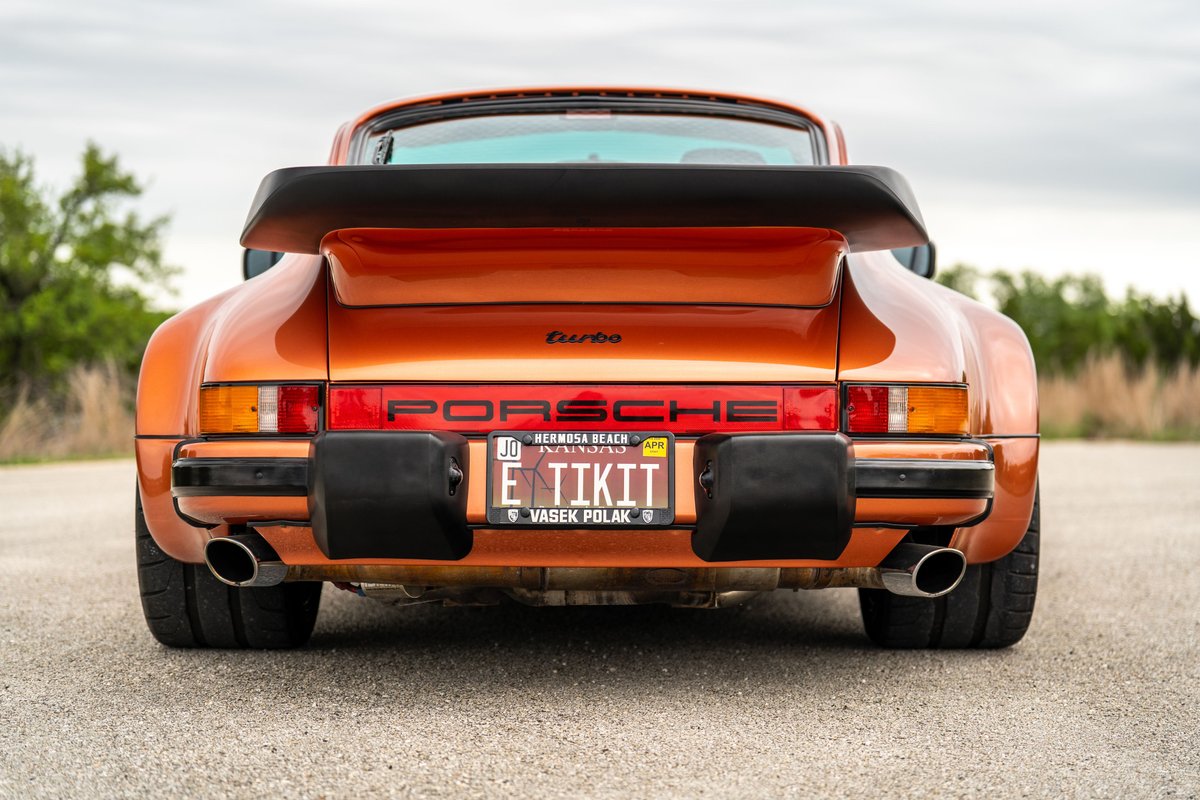 Rear view of an orange metallic 1986 Porsche 930 Turbo with a black whale tail spoiler and dual exhaust pipes.