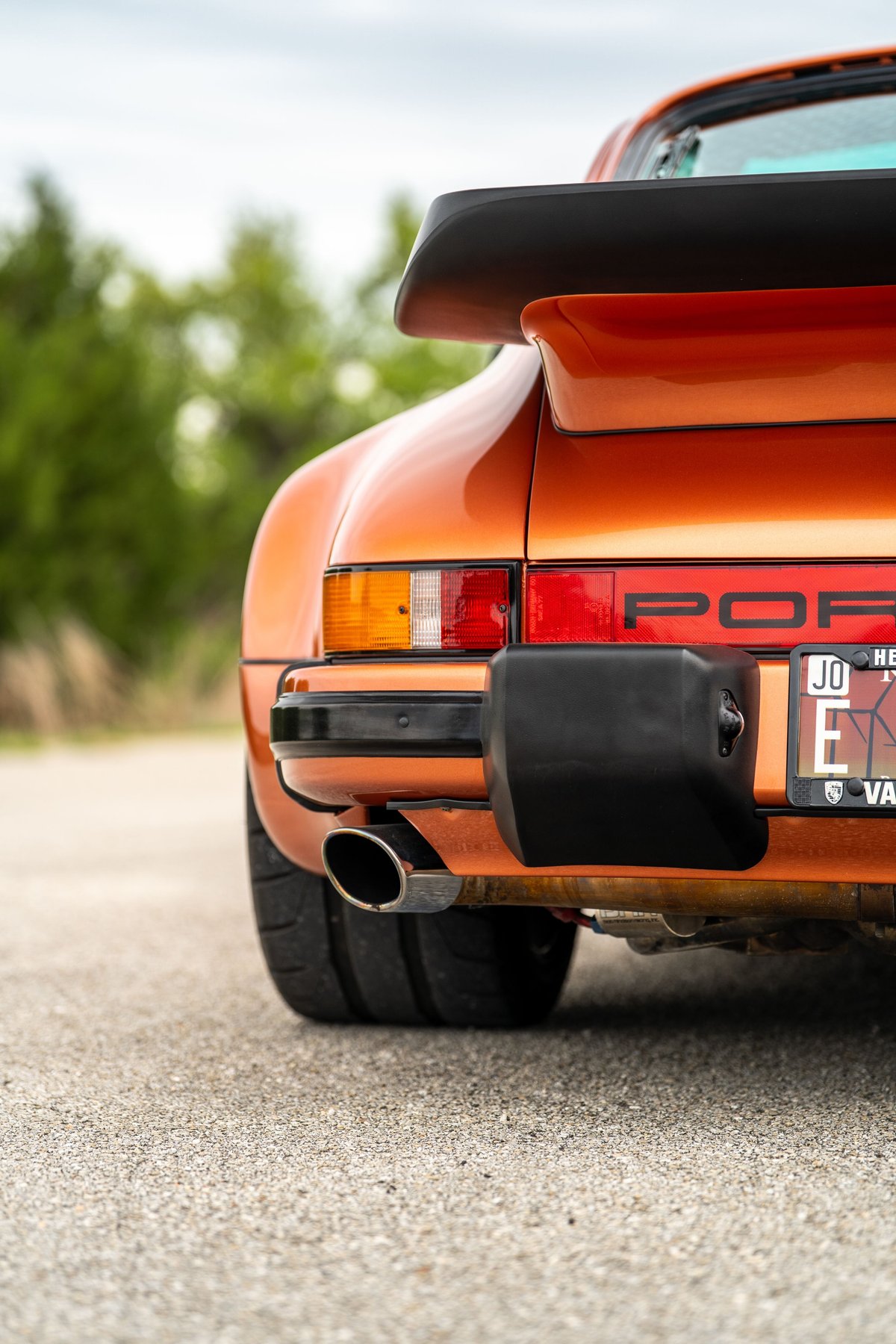 Rear detail of an orange metallic 1986 Porsche 930 Turbo with black whale tail spoiler and chrome exhaust tip.