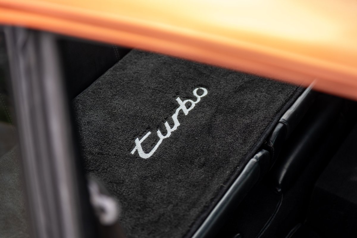 Black floor mat with white embroidered "Turbo" script inside a 1986 orange metallic Porsche 930.