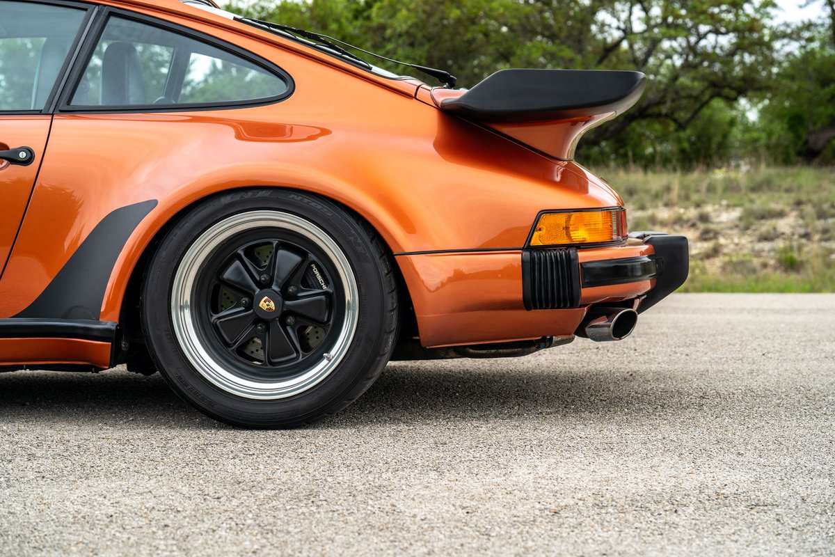 Rear quarter panel of an orange metallic 1986 Porsche 930 Turbo, showing Fuchs rims and whale tail spoiler.