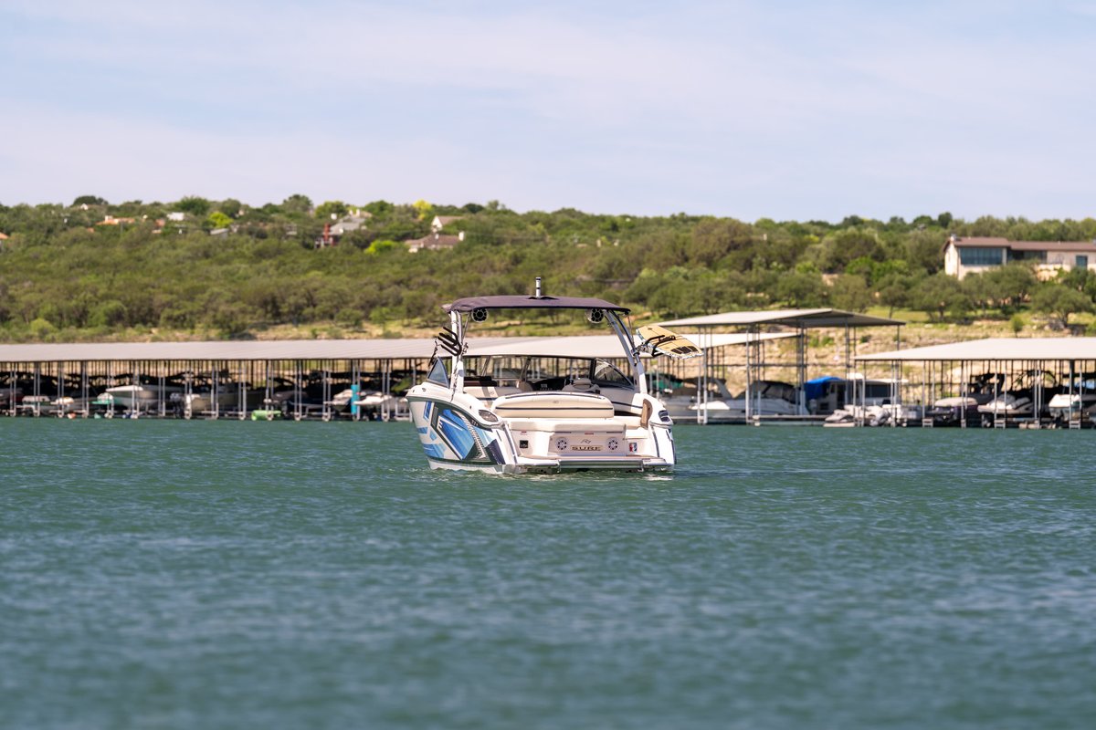 Rear view of a white Cobalt R7 Surf boat with blue livery, cruising on a lake near a marina.