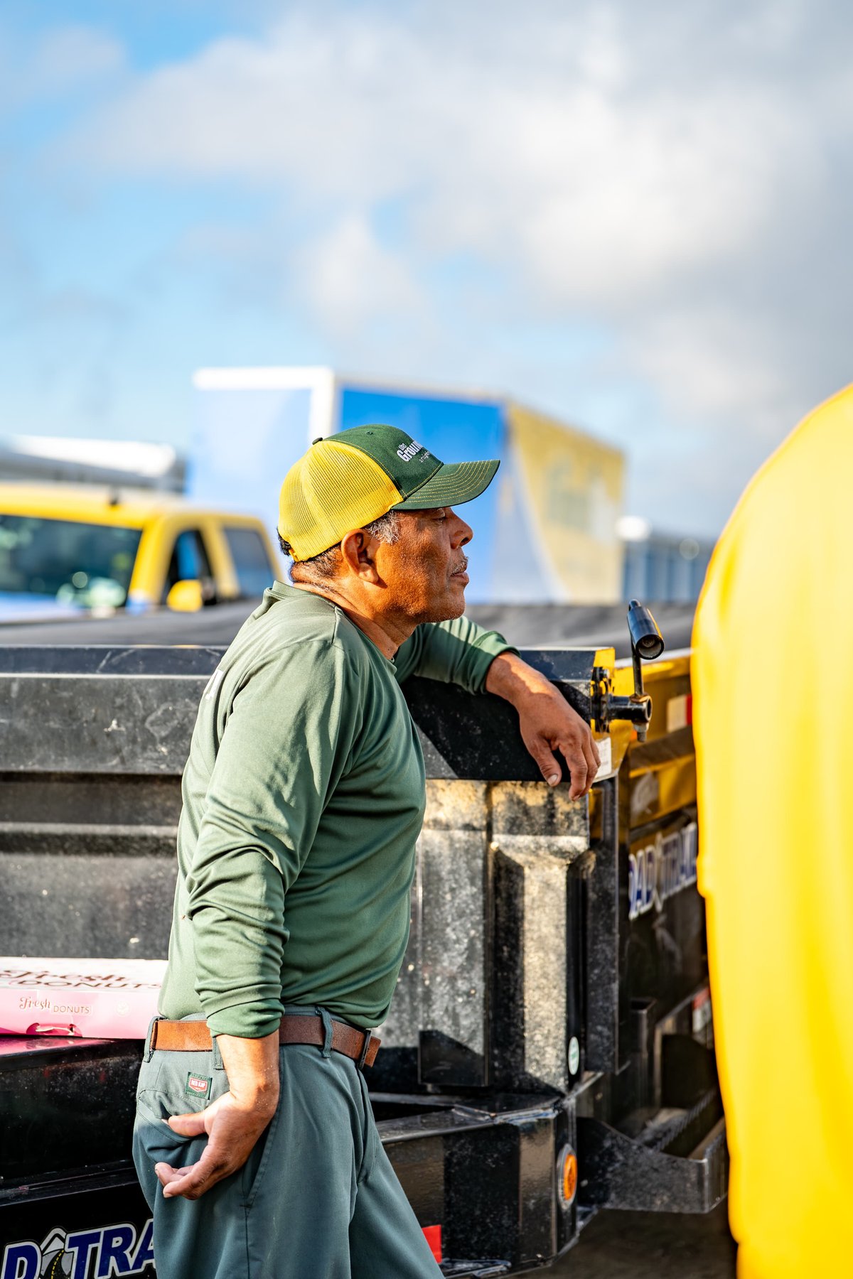 Man in green shirt & cap leans on a black utility trailer, with a yellow pickup truck in the background.