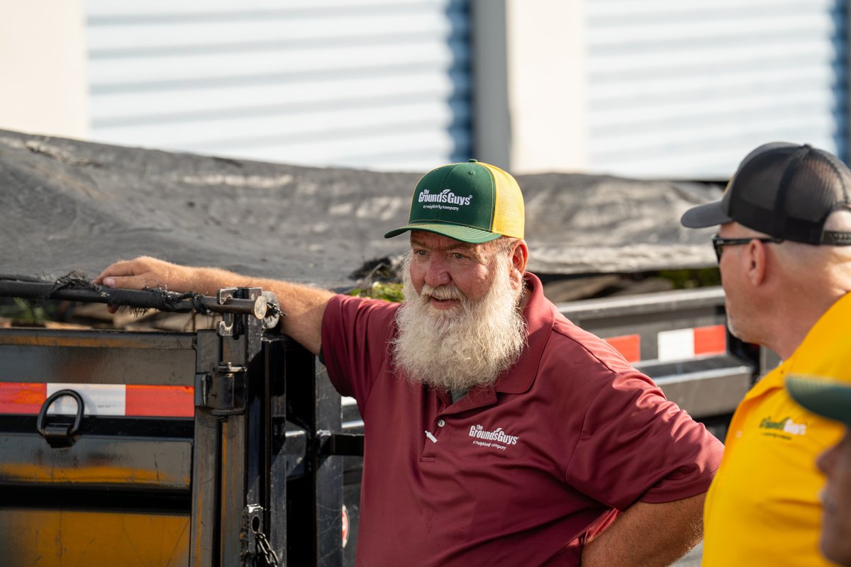 A Grounds Guys employee in a maroon polo leans on a black trailer.