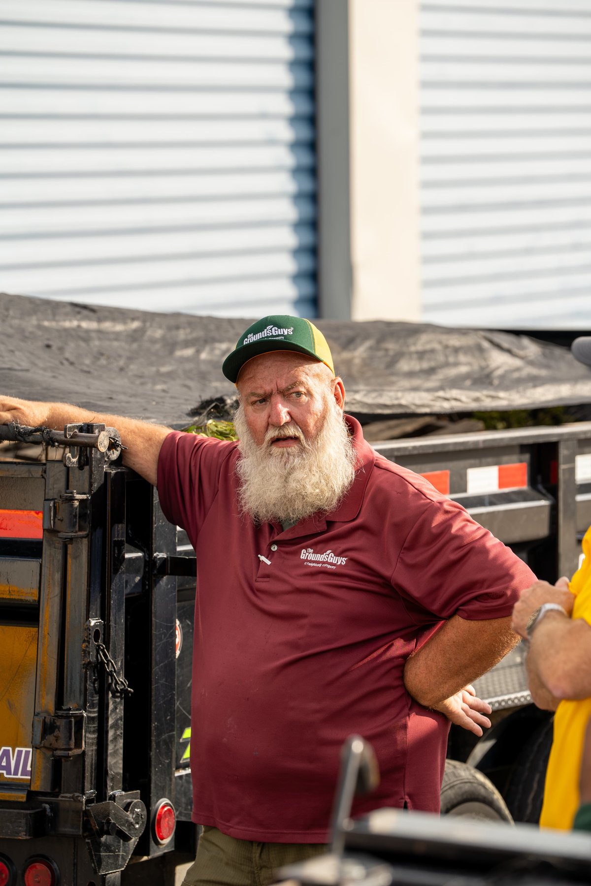 A man in a maroon polo and green cap stands beside a black utility trailer.