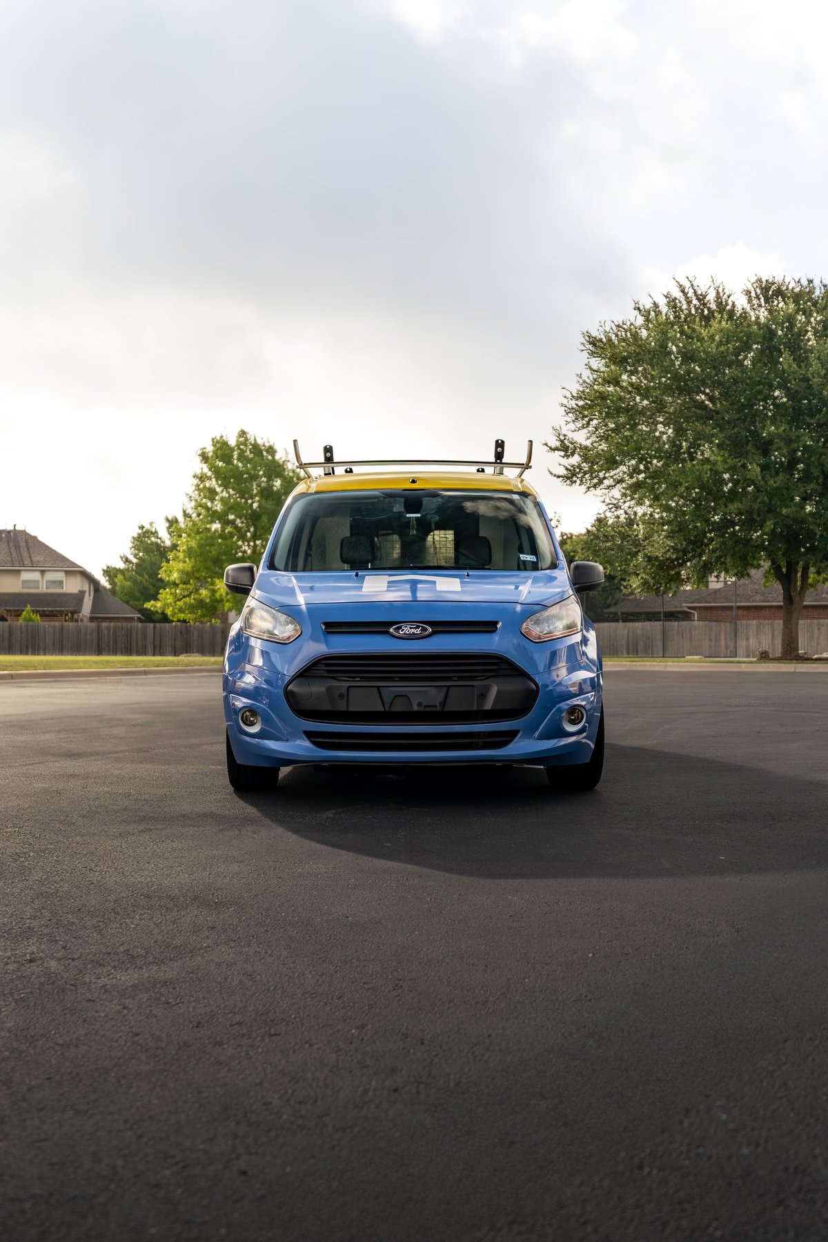 Blue Ford Transit Connect van with yellow roof rack and custom livery, parked outdoors.