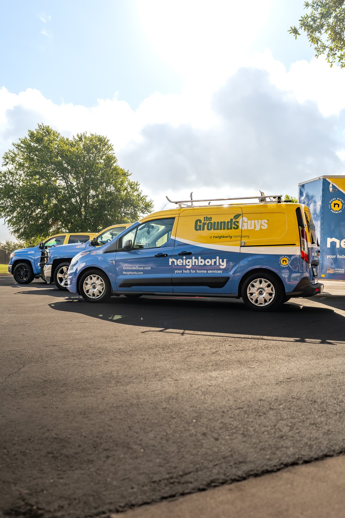 Blue Ford Transit Connect van with yellow "The Grounds Guys" livery and silver rims parked next to a blue pickup truck.