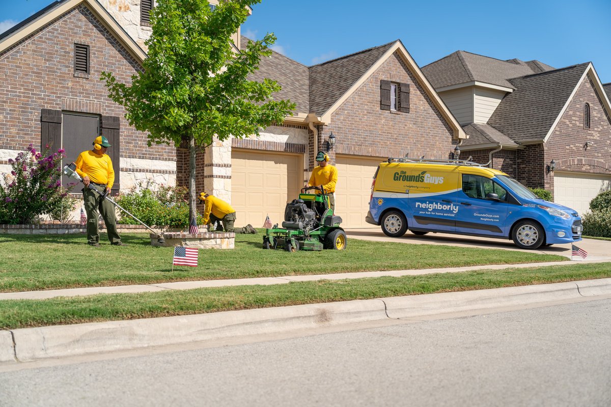 Blue and yellow Ford Transit Connect van with "Grounds Guys" livery, performing lawn maintenance.