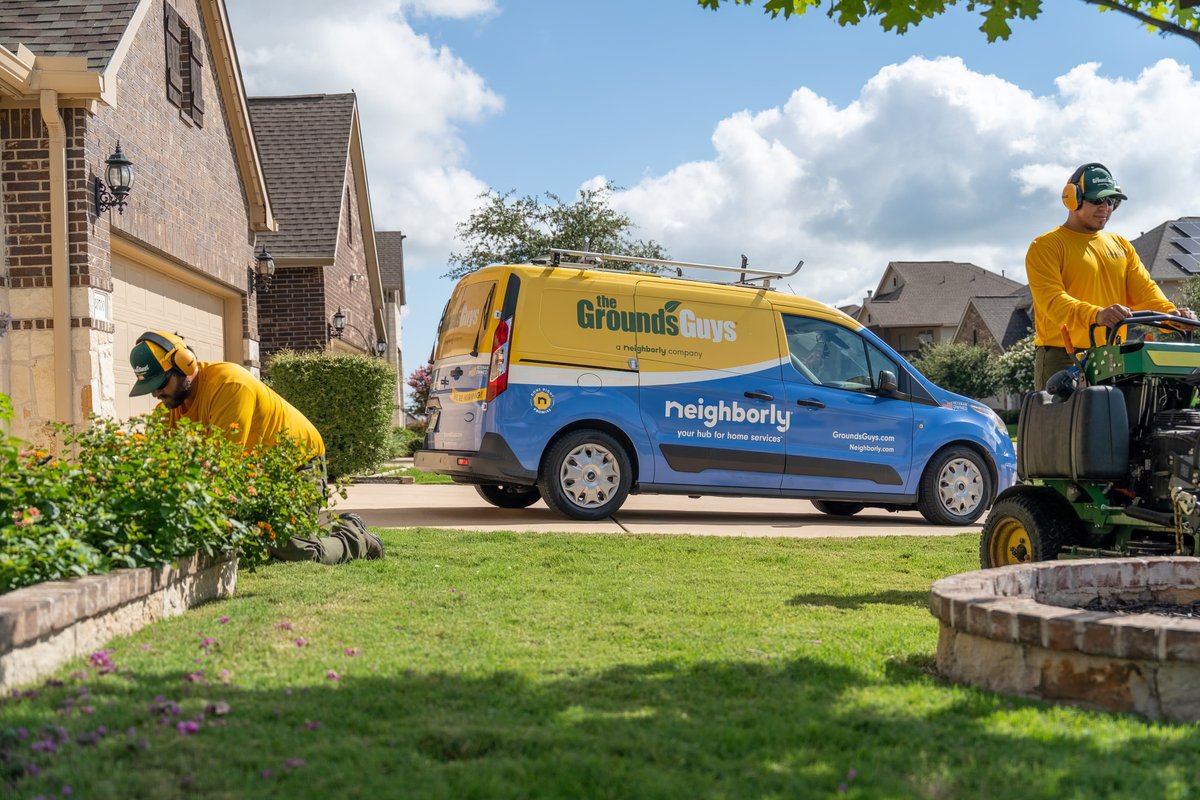 A blue and yellow Ford Transit Connect van with "The Grounds Guys" livery is parked on a suburban street.