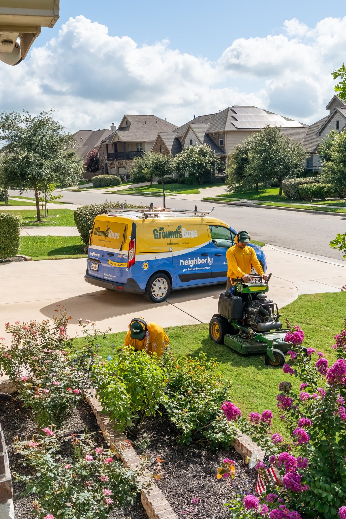 Yellow and blue Grounds Guys van with livery parked on driveway while crew mows lawn.