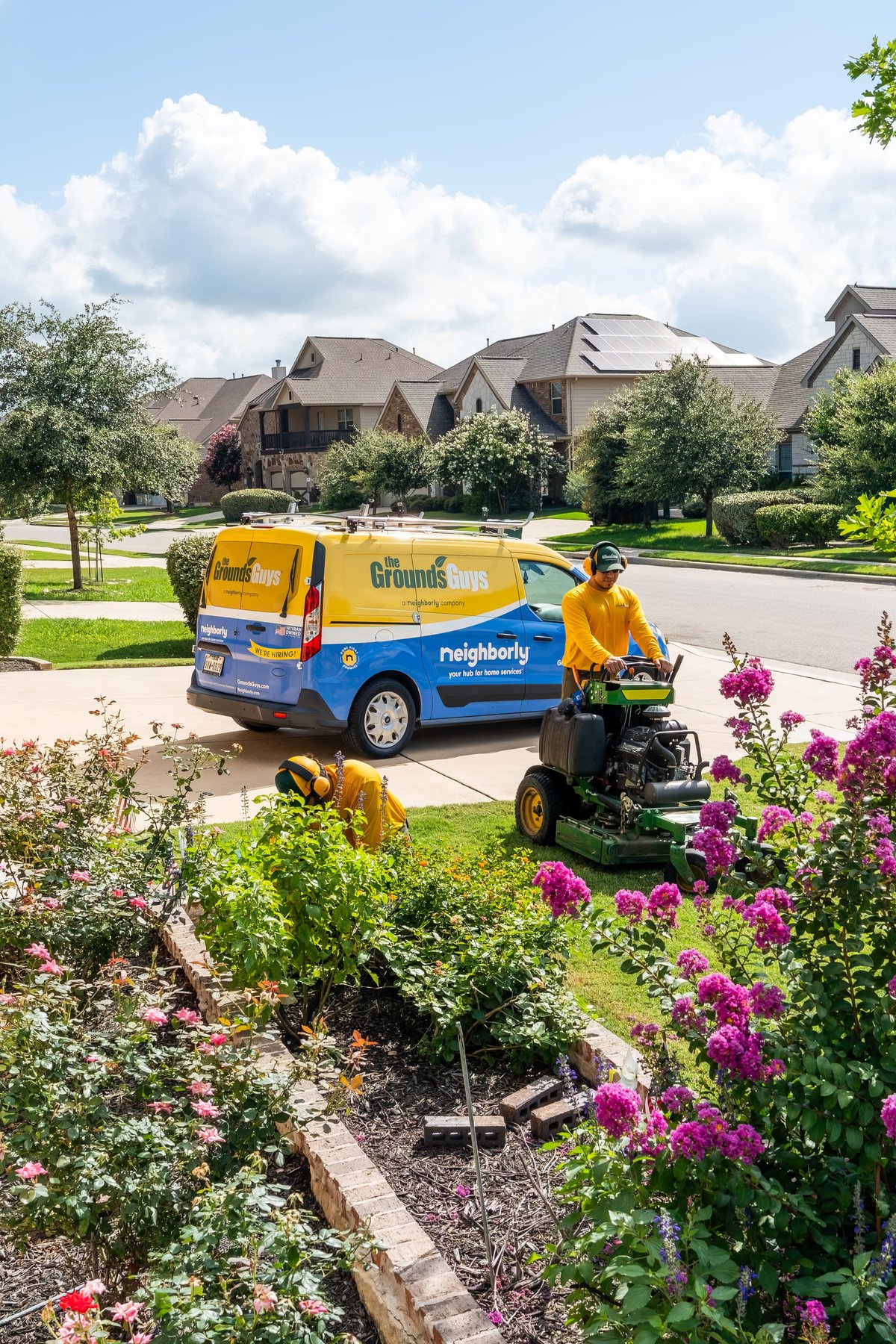 A blue and yellow 2021 Ford Transit Connect with custom "The Grounds Guys" livery and a rider mower in front.