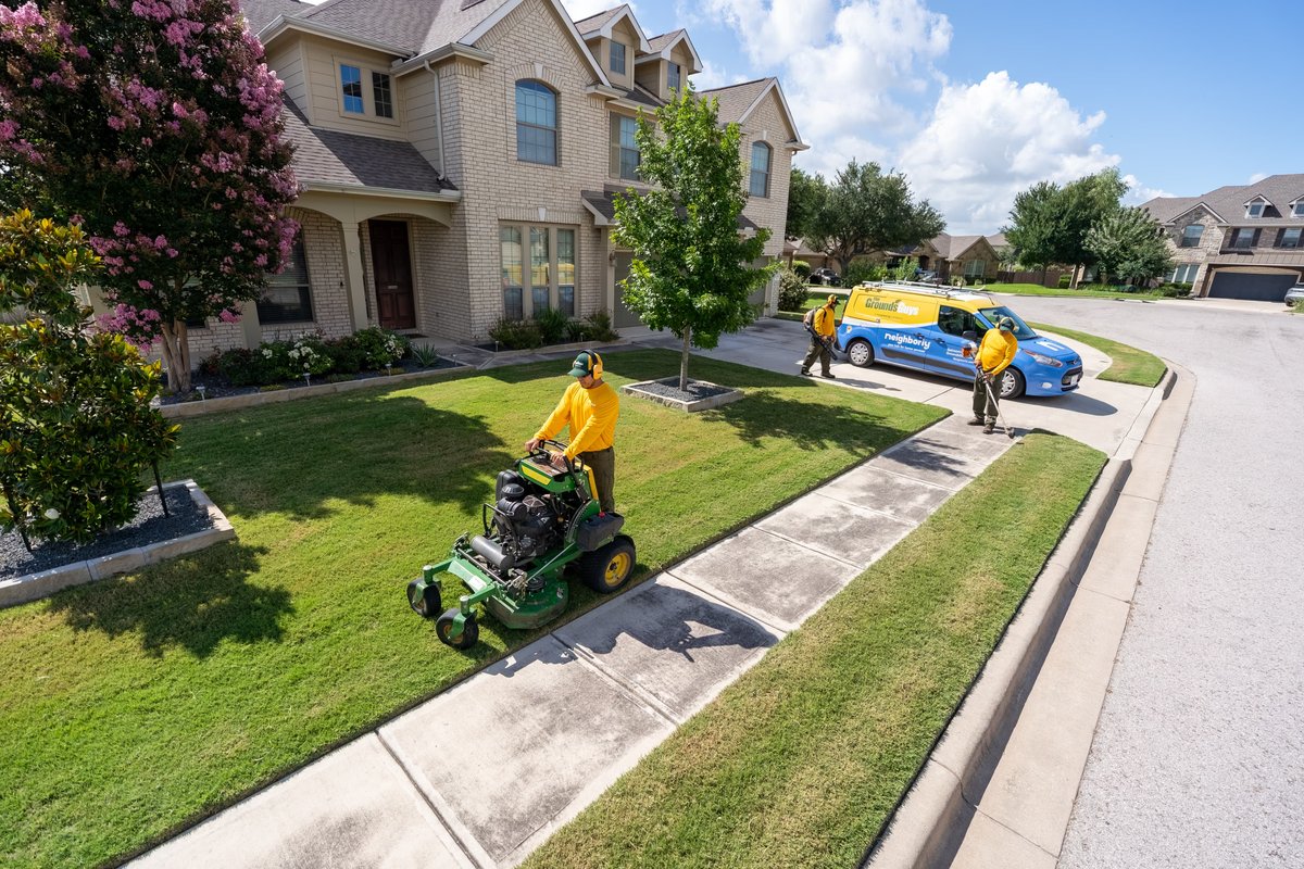 Grounds Guys commercial van in blue with yellow livery, parked by a manicured lawn and house.
