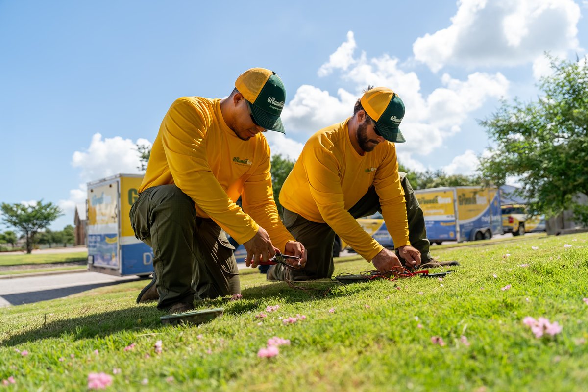 Two men in yellow shirts work on wires in the grass, near a yellow and blue trailer.