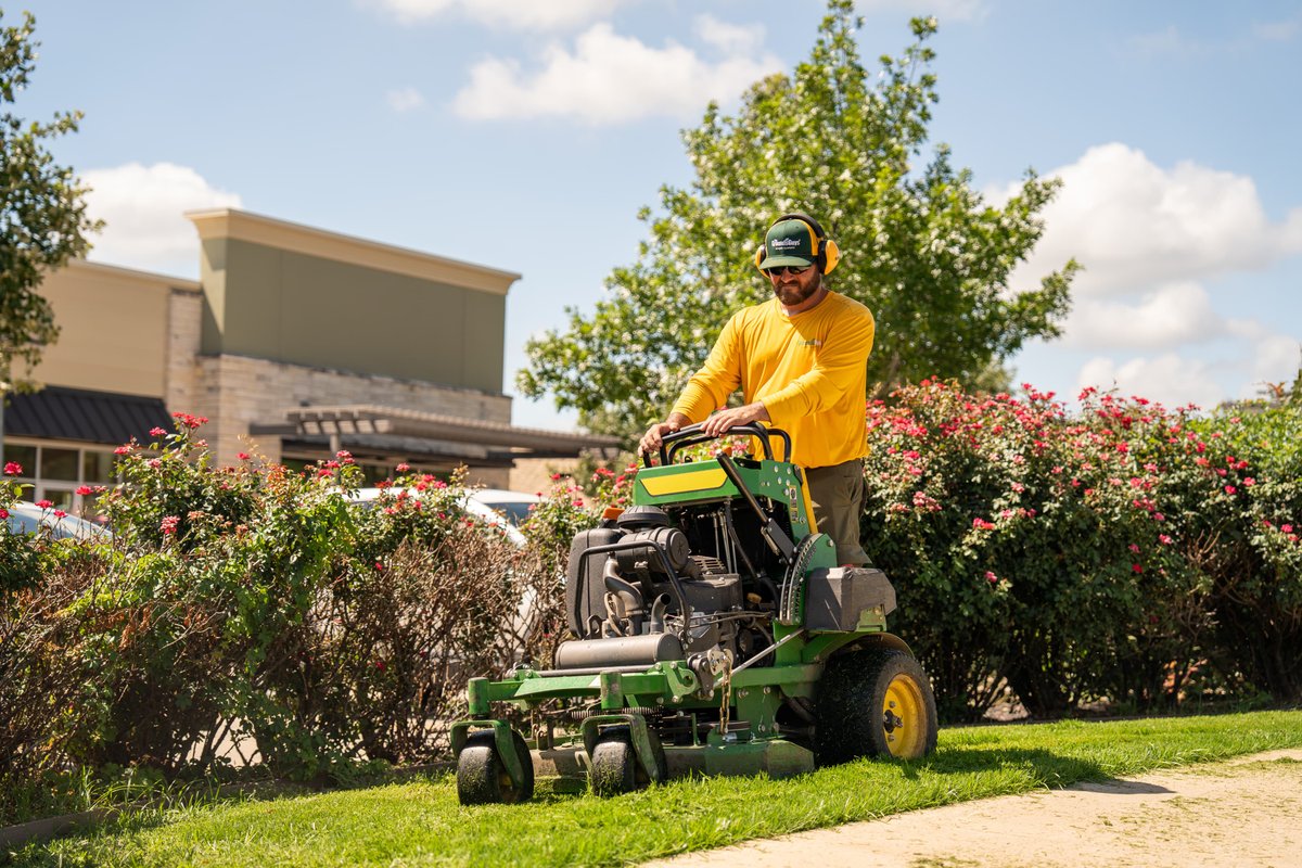 Green John Deere zero-turn mower, yellow rims, driving on grass.