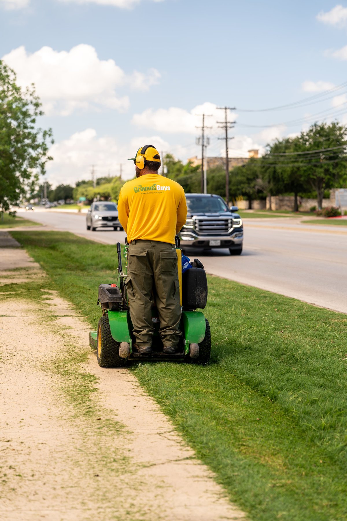 Man on green zero-turn mower with yellow "Grounds Guys" livery, driving on roadside.