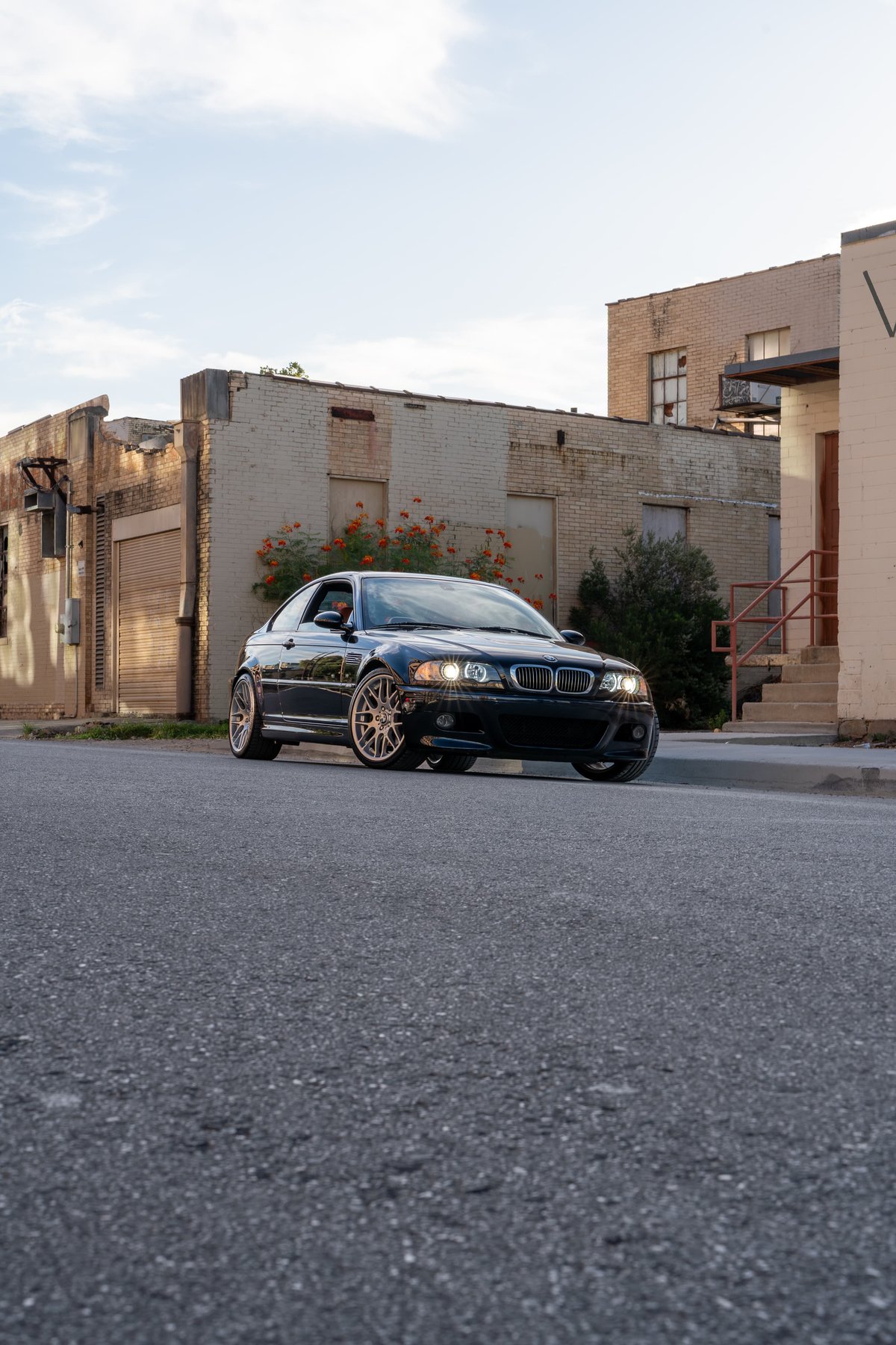 Black 2002 BMW M3 Coupe with gold rims parked on asphalt, with urban buildings in background.