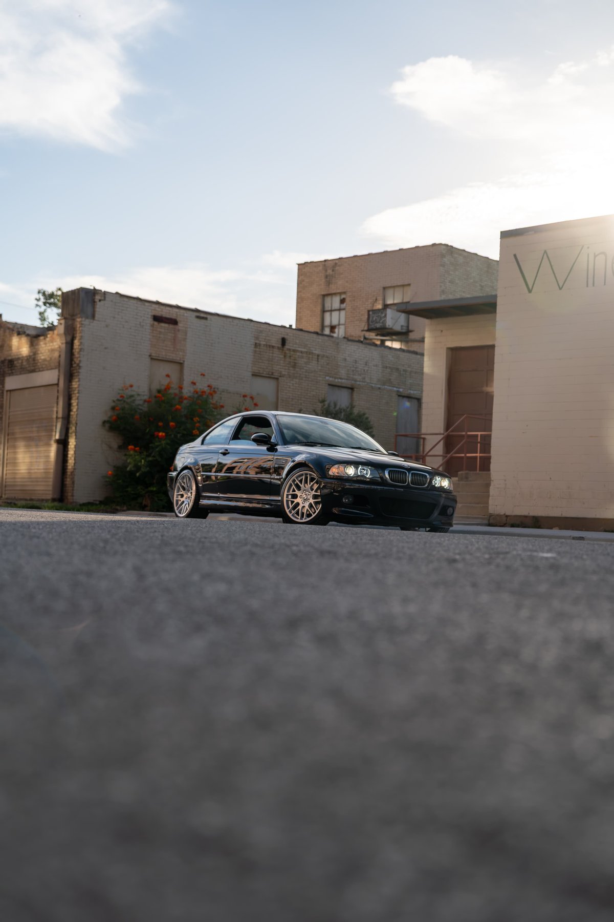 Black 2002 BMW M3 coupe on gold multi-spoke rims parked near buildings.