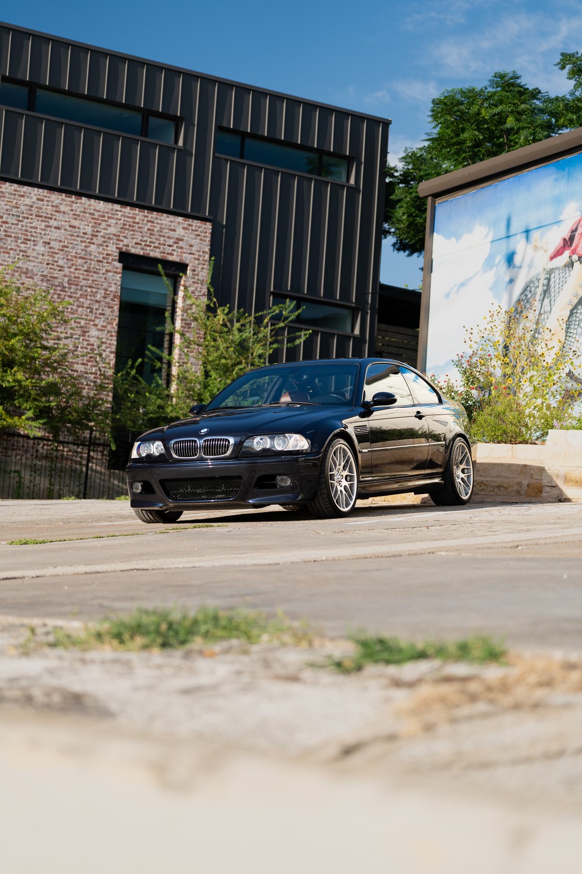 Black 2002 BMW M3 coupe with silver multi-spoke rims parked in front of a modern building.