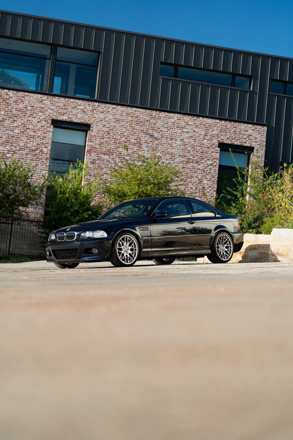 Black 2002 BMW M3 coupe with silver rims parked in front of a modern building.