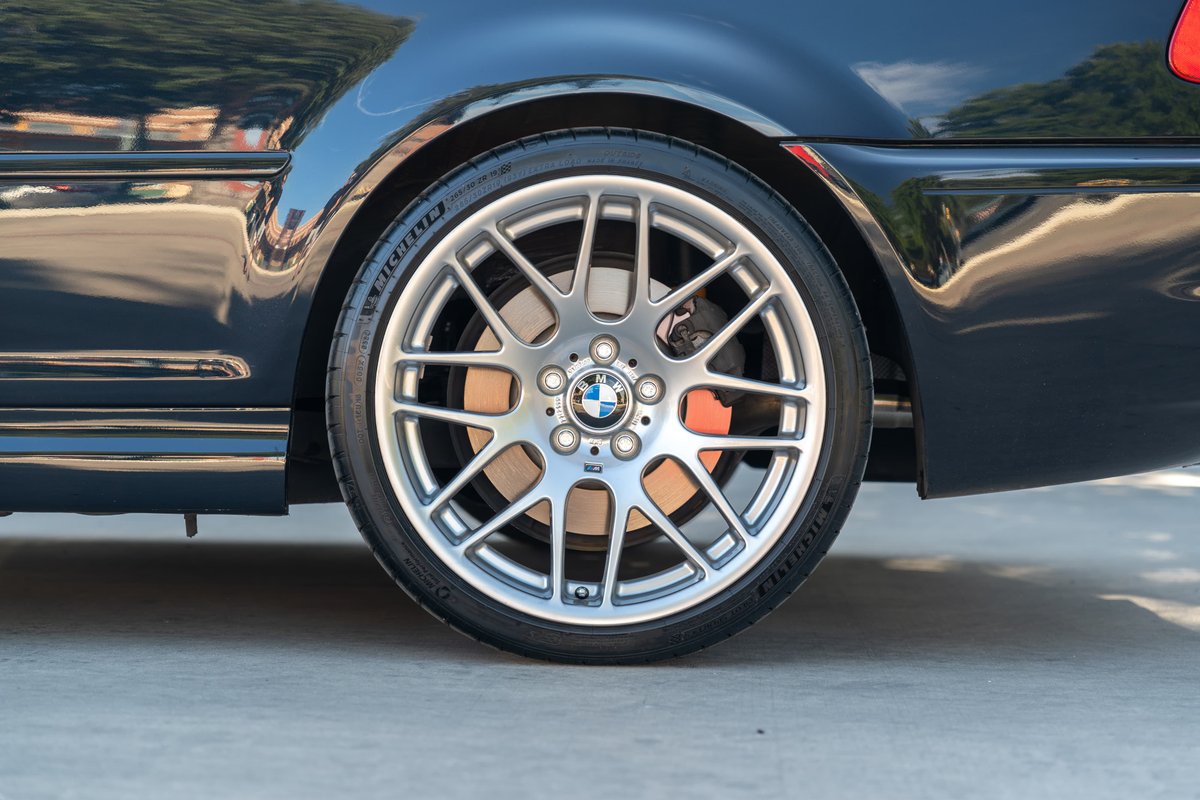 Close-up of a carbon black 2002 BMW M3 rear wheel with silver alloy rims and orange brake calipers.