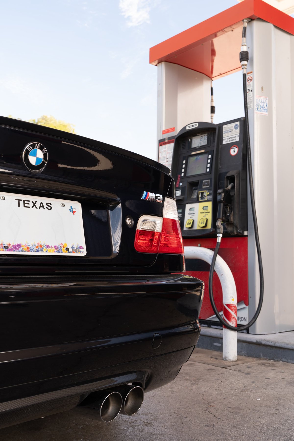 Rear view of a black 2002 BMW M3 at a gas pump, featuring dual exhaust tips and M3 badging.