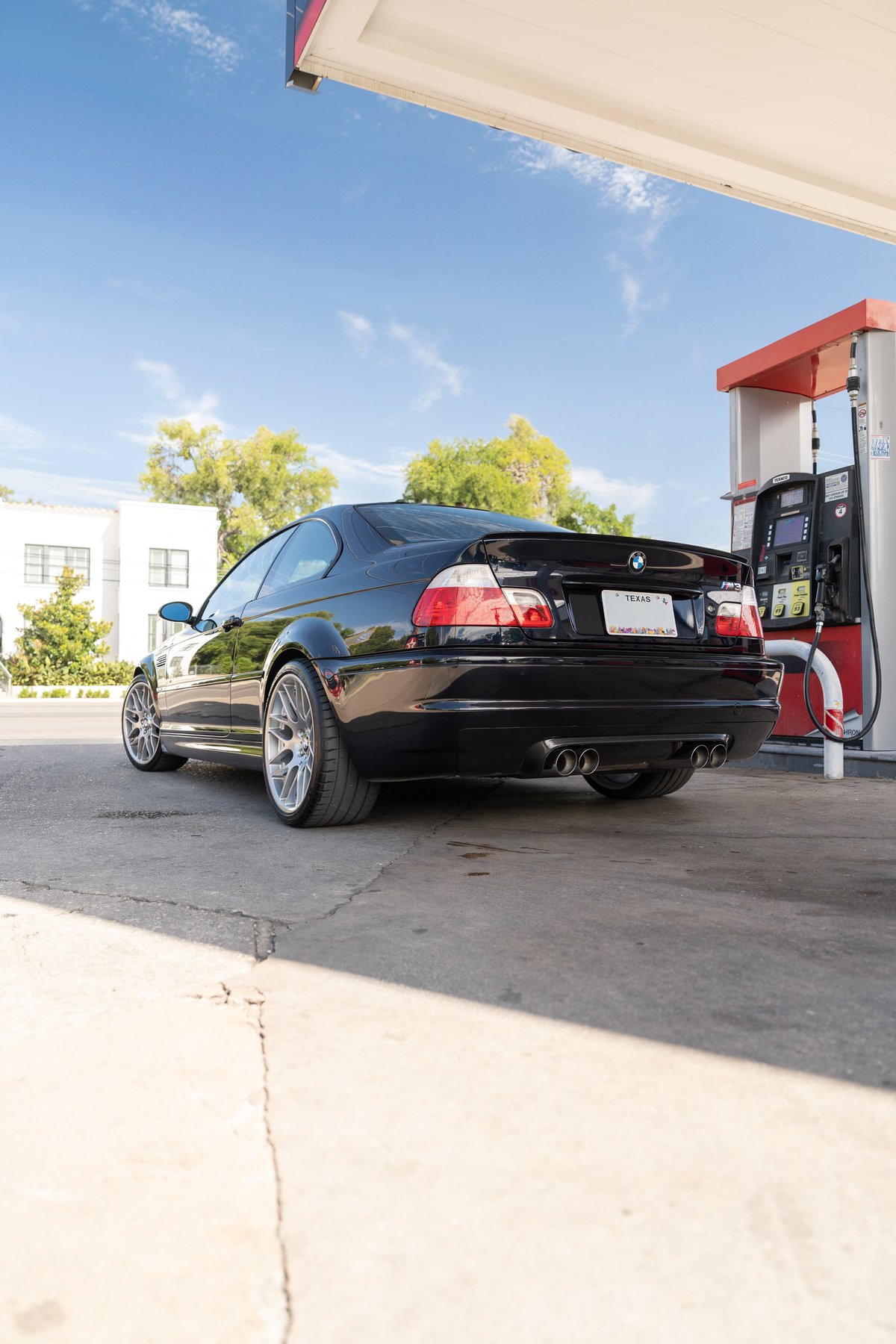 Black 2002 BMW M3 E46 coupe at gas pump, showing quad exhaust, rear spoiler, and silver rims.